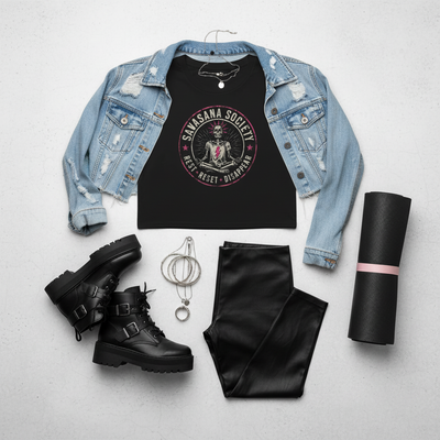 A styled flat-lay outfit on a light gray background featuring a black graphic tank top with a 'Samsara Society' print, layered under a light-wash distressed denim jacket. The outfit is accessorized with black combat boots, black faux-leather pants, a silver chain necklace, and a rolled-up black yoga mat with a pink band.