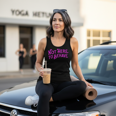 A woman wearing a black tank top with the text 'NOT HERE TO BEHAVE' in pink sits cross-legged on the hood of a black car, holding an iced coffee and a yoga mat. A blurred building with 'YOGA HETU.