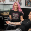 A woman with pink hair sits in a tattoo chair while another person tattoos her arm. The seated woman wears a black graphic T-shirt with the phrase 'ZEN AS HELL' in white and pink lettering with a snake design.