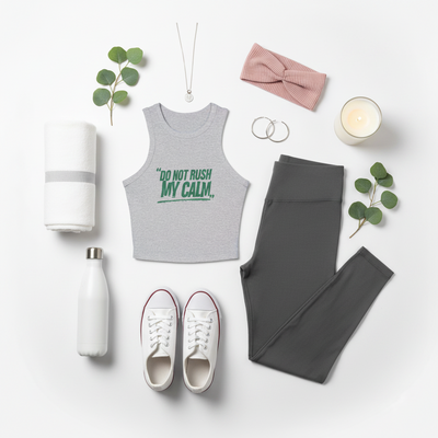 A styled flat lay of activewear and wellness accessories on a white background, featuring a heather gray ribbed tank top with the green text 'DO NOT RUSH MY CALM.', dark gray leggings, white sneakers, a white water bottle, a white towel, a pink bow headband, silver hoop earrings, a necklace, and green eucalyptus sprigs.