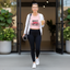 A woman walks out of a building wearing a pink graphic tank top that reads 'NAMASTE BITCHES' with a red lotus design, black leggings, a white zip-up cardigan, and white sneakers. She carries a yoga mat and holds an iced coffee.