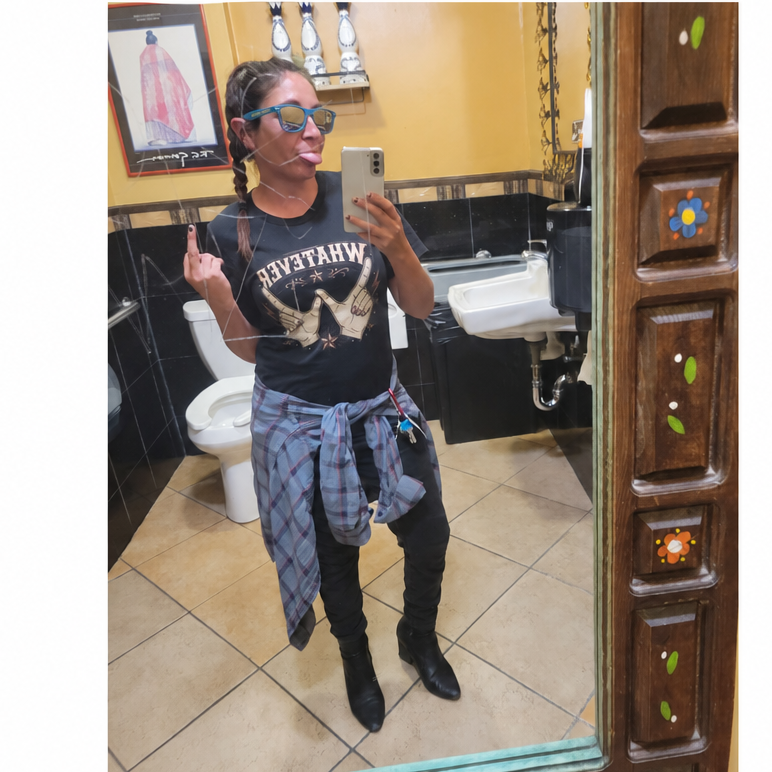 A woman taking a mirror selfie in a bathroom, wearing a black graphic t-shirt with 'WHATEVER', a blue plaid shirt tied around her waist, black pants, and black ankle boots.