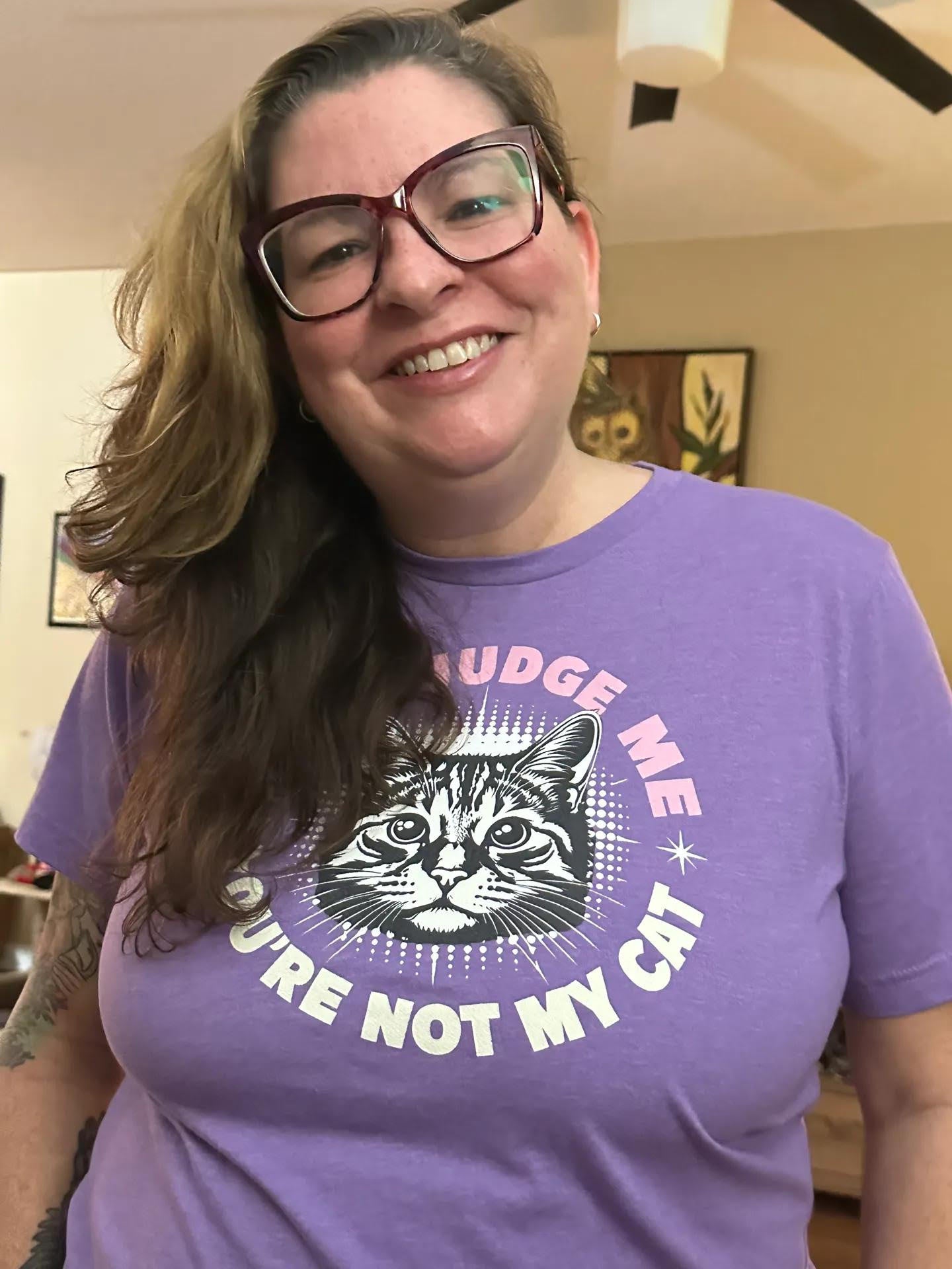 A smiling woman with glasses wearing a purple graphic t-shirt that reads 'JUDGE ME YOU'RE NOT MY CAT' with a black and white cat illustration.