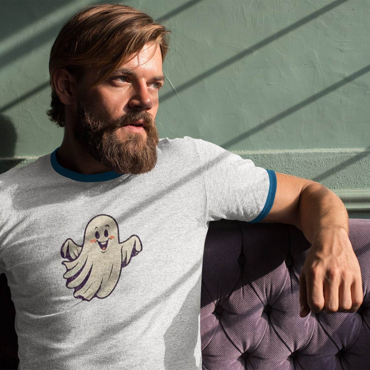 A man with a beard wearing a light gray ringer t-shirt with a cartoon ghost graphic and blue trim, sitting on a purple tufted couch against a green wall with window shadow patterns.
