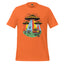 An orange short-sleeve crewneck t-shirt featuring a colorful retro graphic of three UFOs with rainbow beams, a yellow car, and cows on a grassy field, displayed against a solid white background.